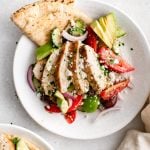 Overhead view of sliced chicken breast on Greek salad with pita and avocado.