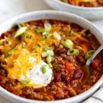 White shallow bowl filled with the best chili recipe and topped with shredded cheddar cheese, sour cream, and green onions.