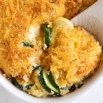 Wooden spoon scooping cheesy zucchini casserole from a white casserole dish.