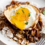White plate filled with fluffy white rice smothered in an onion and mushroom gravy and topped with a hamburger patty and runny fried egg and garnished with sliced green onion.