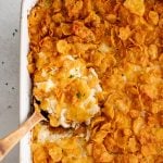 Wooden serving spoon in a large white casserole dish filled with gooey, cheesy baked funeral potatoes topped with crispy cornflakes.