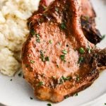 Two beautifully seasoned bone-in pork chops tilted against a pile of mashed potatoes on a white serving plate garnished with chopped parsley.